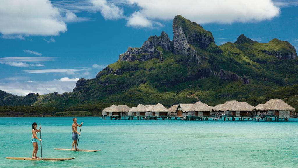 Couple paddleboarding in front of Four Seasons Bora Bora overwater bungalows with Mt. Otemanu